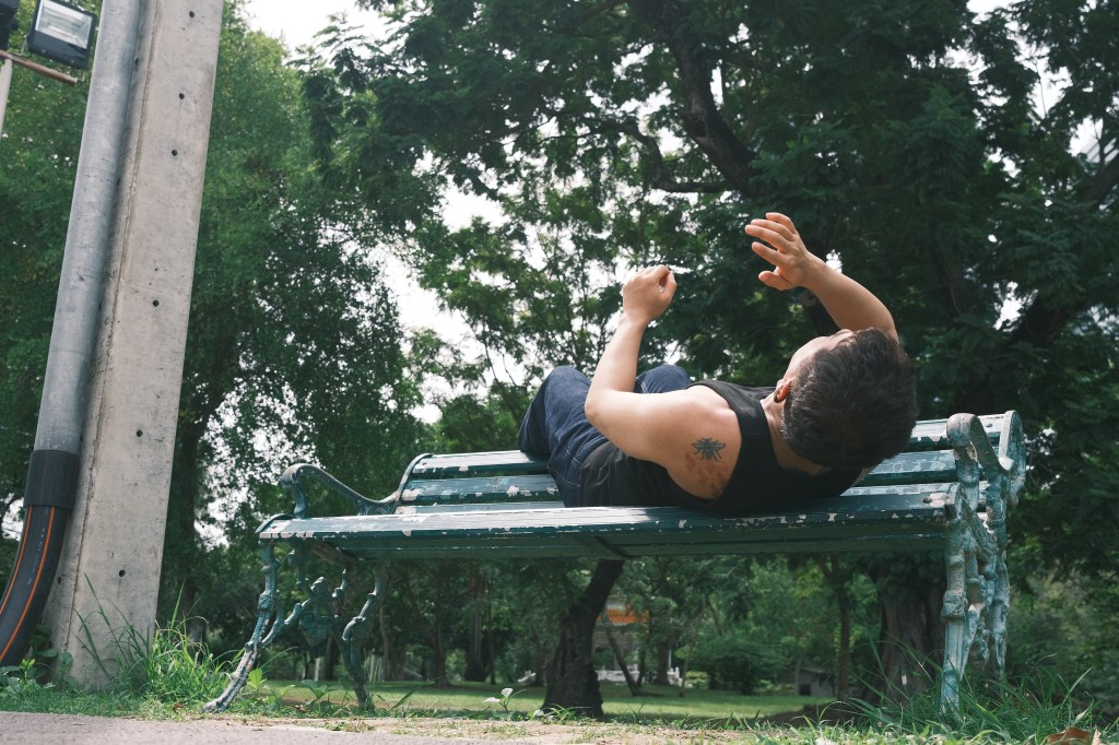 Harris reclines on a park bench, raising his hands beneath the trees.